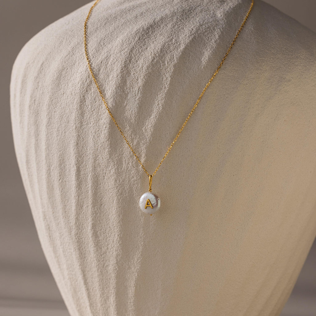 The Pearl Initial Pendant Necklace features a round white pendant with the letter A, displayed on a textured beige stand—a beautiful personalized gift perfect for any occasion.