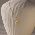 The Pearl Initial Pendant Necklace features a round white pendant with the letter A, displayed on a textured beige stand—a beautiful personalized gift perfect for any occasion.