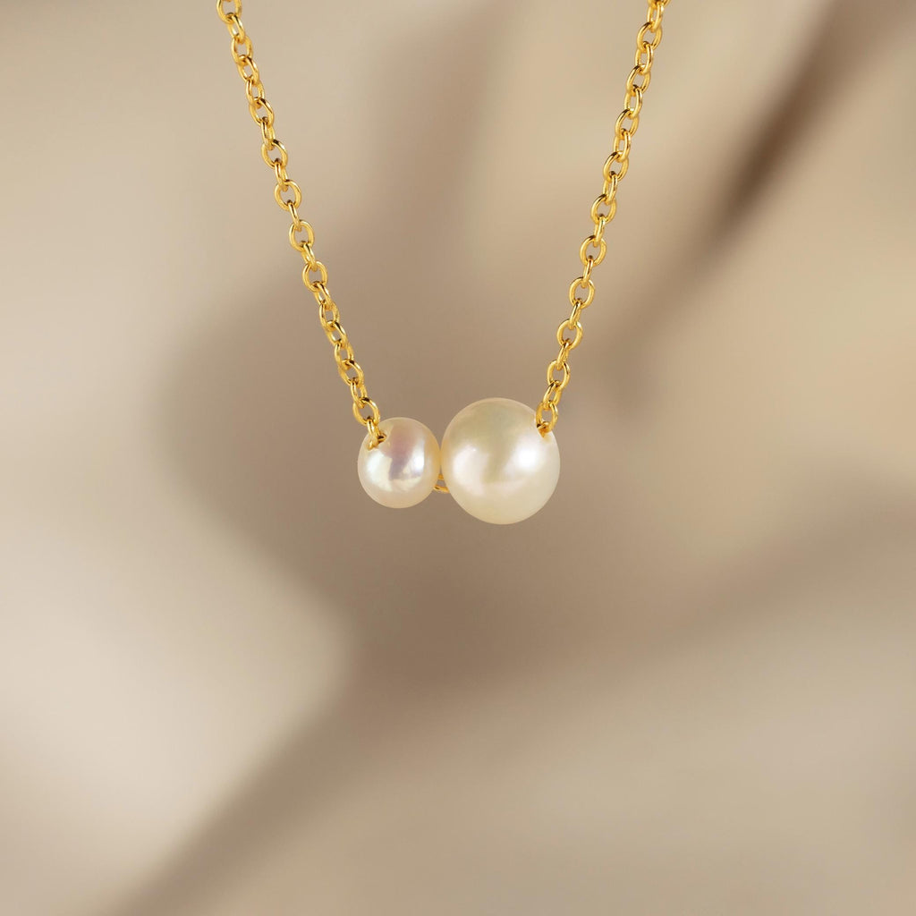 The Dainty Duo Pearl Necklace features two white pearls of varying sizes on a gold chain, set against a soft beige background—an elegant choice for a wedding gift.