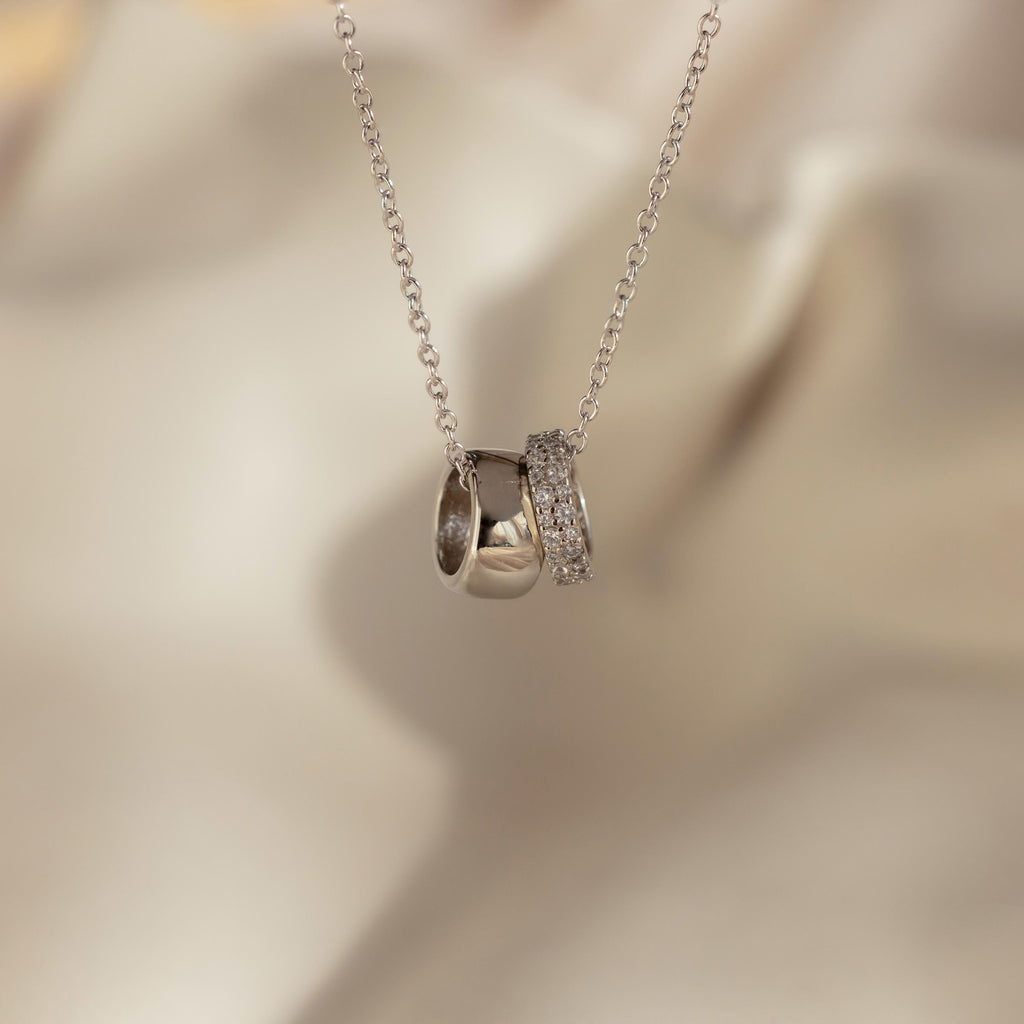 The Pave Duo Circle Necklace, featuring two rings—one plain and one pavé-set with sparkling stones—rests elegantly on a soft beige background.