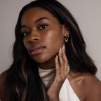 A woman with long, wavy hair and a cream top poses with her hand near her face, highlighting the Bold Teardrop Hoops gold earrings.