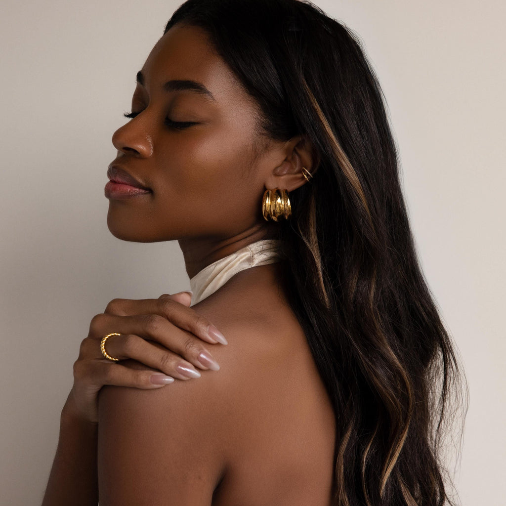 A woman with long dark hair and gold jewelry poses with eyes closed against a plain background, highlighting the Chunky Triple Hoops that make a bold statement.