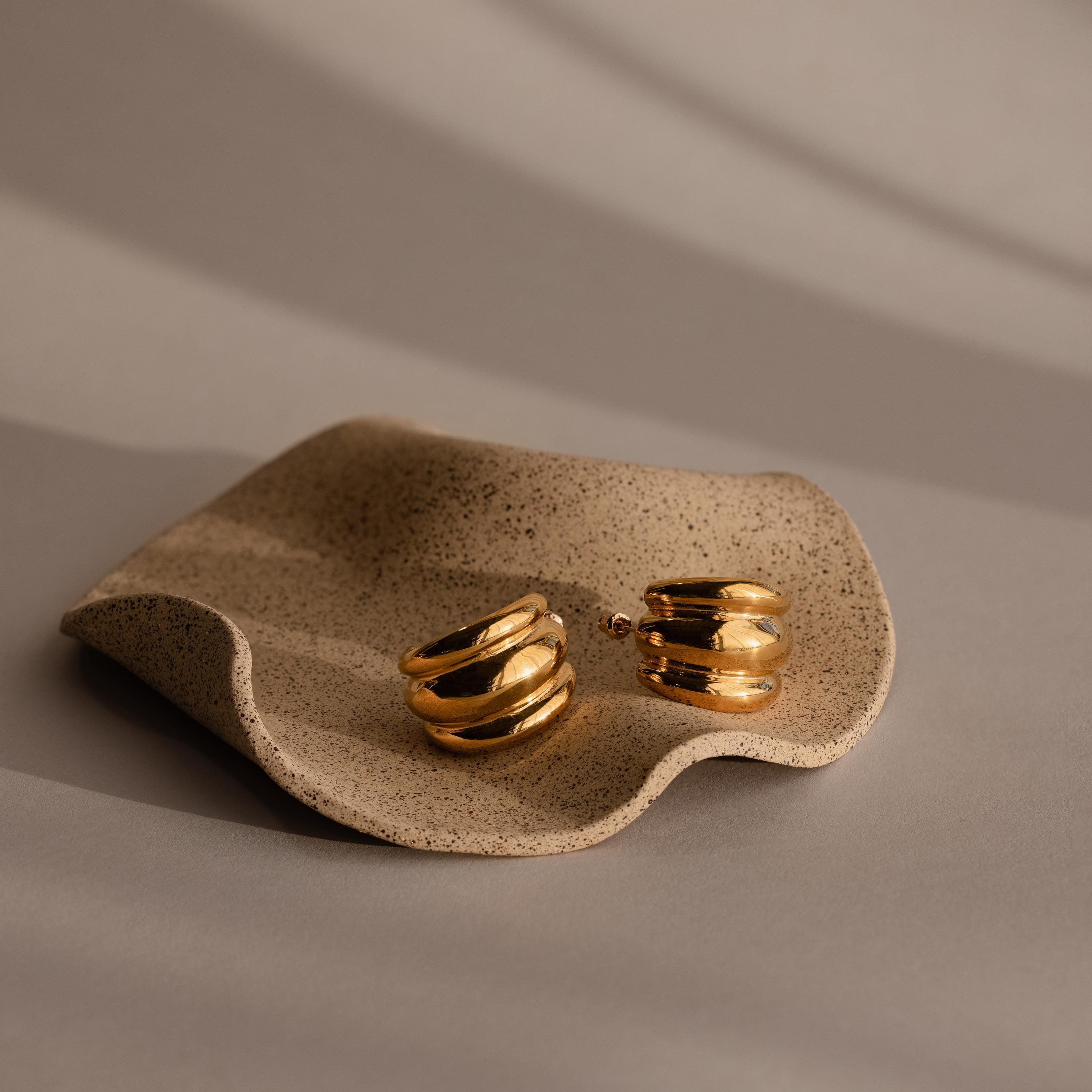 The Chunky Triple Hoops rest on a wavy, textured beige ceramic dish with soft shadows—bold statement earrings to elevate any style.