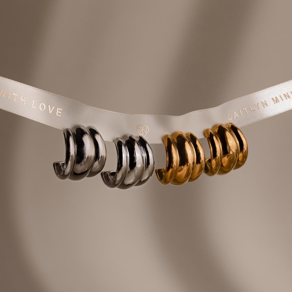 The Chunky Triple Hoops, a bold pair of statement earrings in silver and gold, hang on a branded white ribbon against a neutral background.