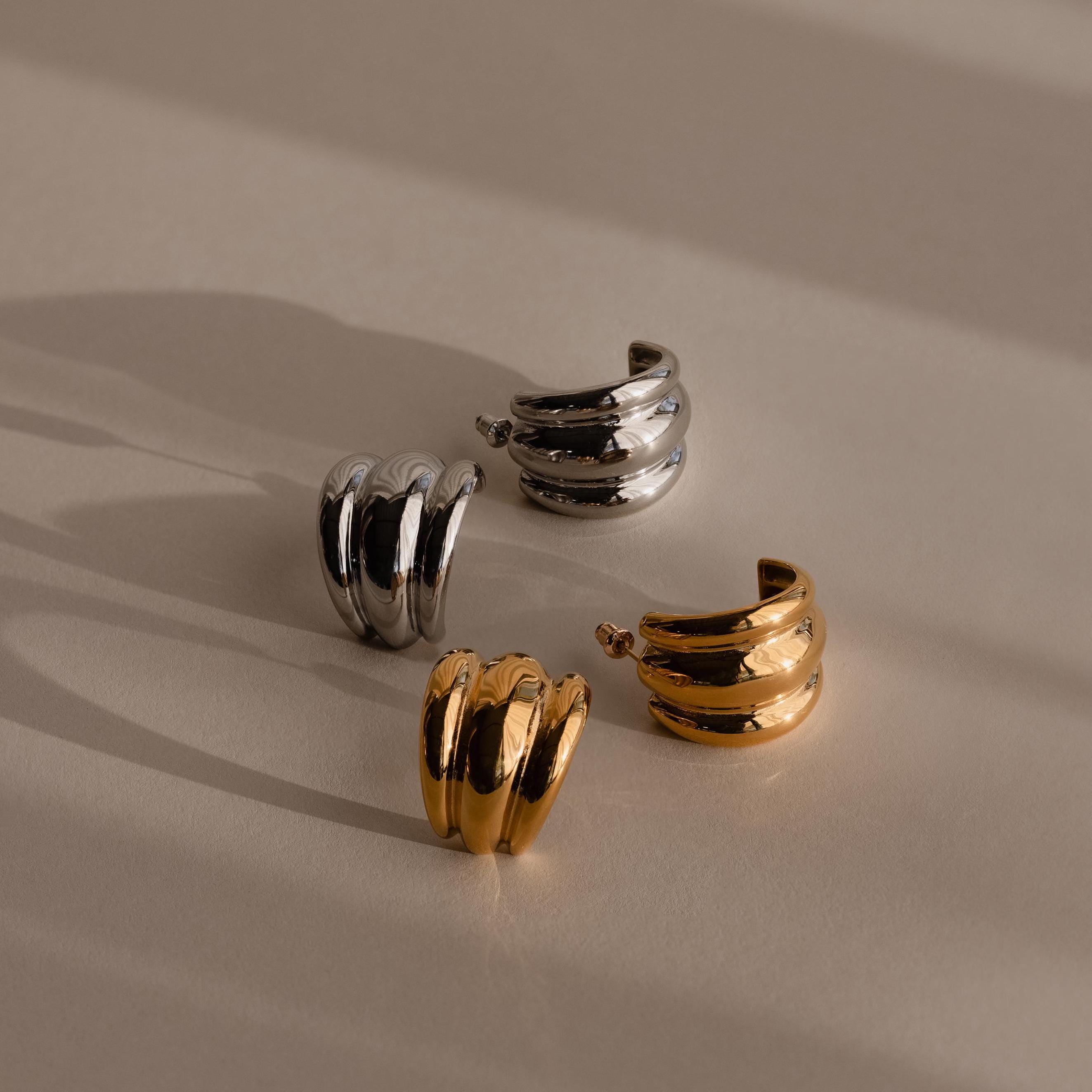 Displayed on a beige surface with soft shadows are two pairs of Chunky Triple Hoops statement earrings in gold and silver.