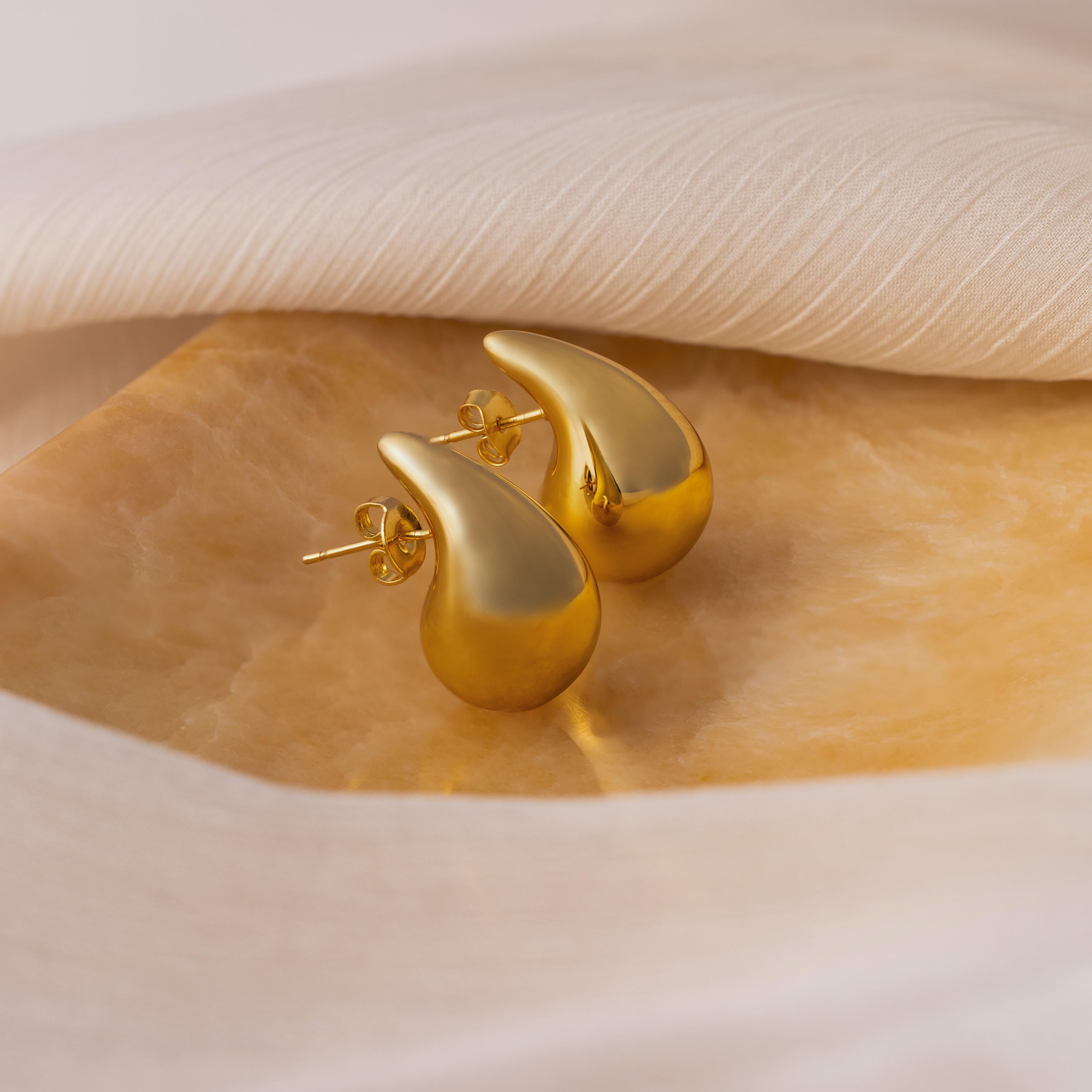 Chunky Tear Drop Studs: Gold teardrop-shaped earrings showcased on a golden surface with soft white fabric, these studs bring effortless elegance to any outfit.