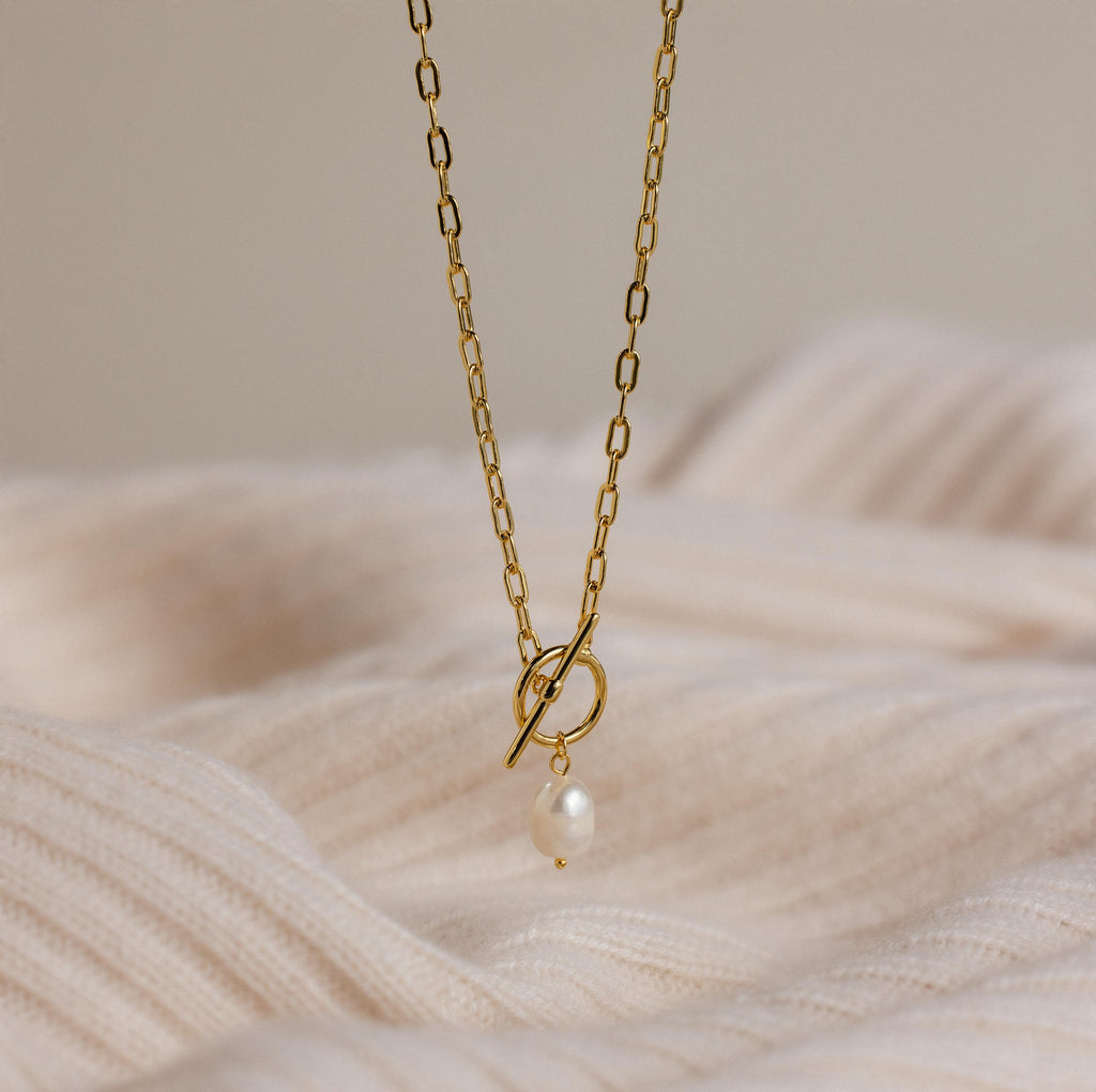 The Paperclip Pearl Toggle Necklace features a gold chain with a single pearl drop and toggle clasp, displayed on soft, cream-colored textured fabric—a timeless pearl jewelry piece.