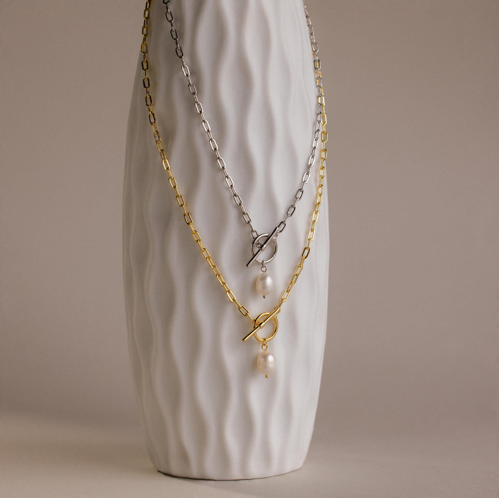 A textured white vase displays two Paperclip Pearl Toggle Necklaces—one gold, one silver—each with pearls and a toggle clasp, making them the perfect statement pearl jewelry pieces.