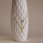 A textured white vase displays two Paperclip Pearl Toggle Necklaces—one gold, one silver—each with pearls and a toggle clasp, making them the perfect statement pearl jewelry pieces.