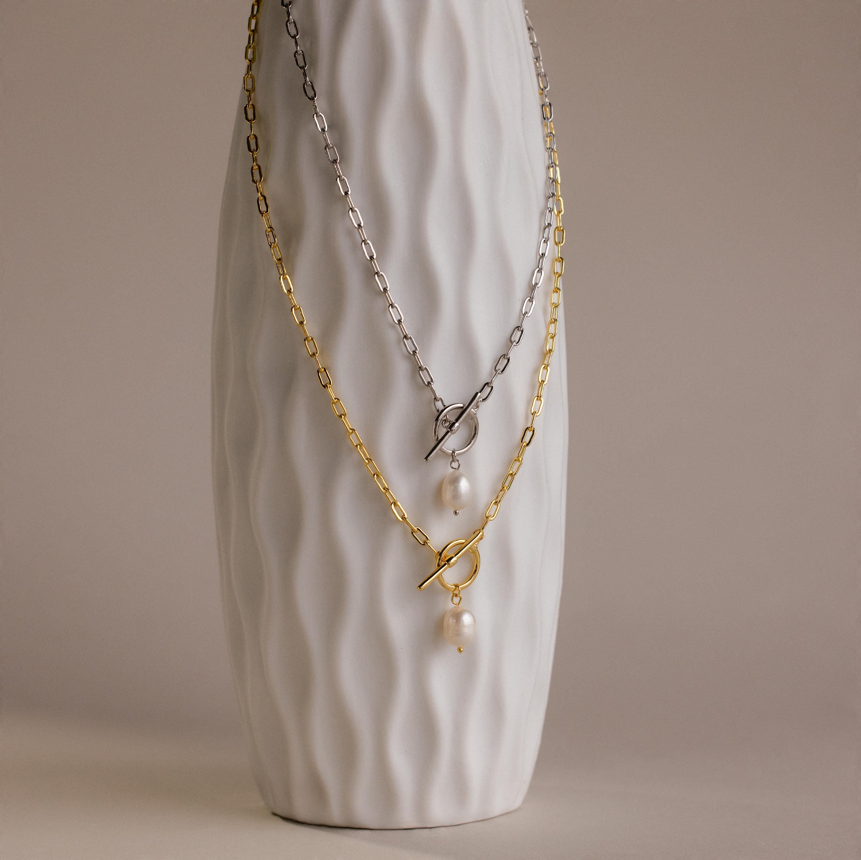 A textured white vase displays two Paperclip Pearl Toggle Necklaces—one gold, one silver—each with pearls and a toggle clasp, making them the perfect statement pearl jewelry pieces.