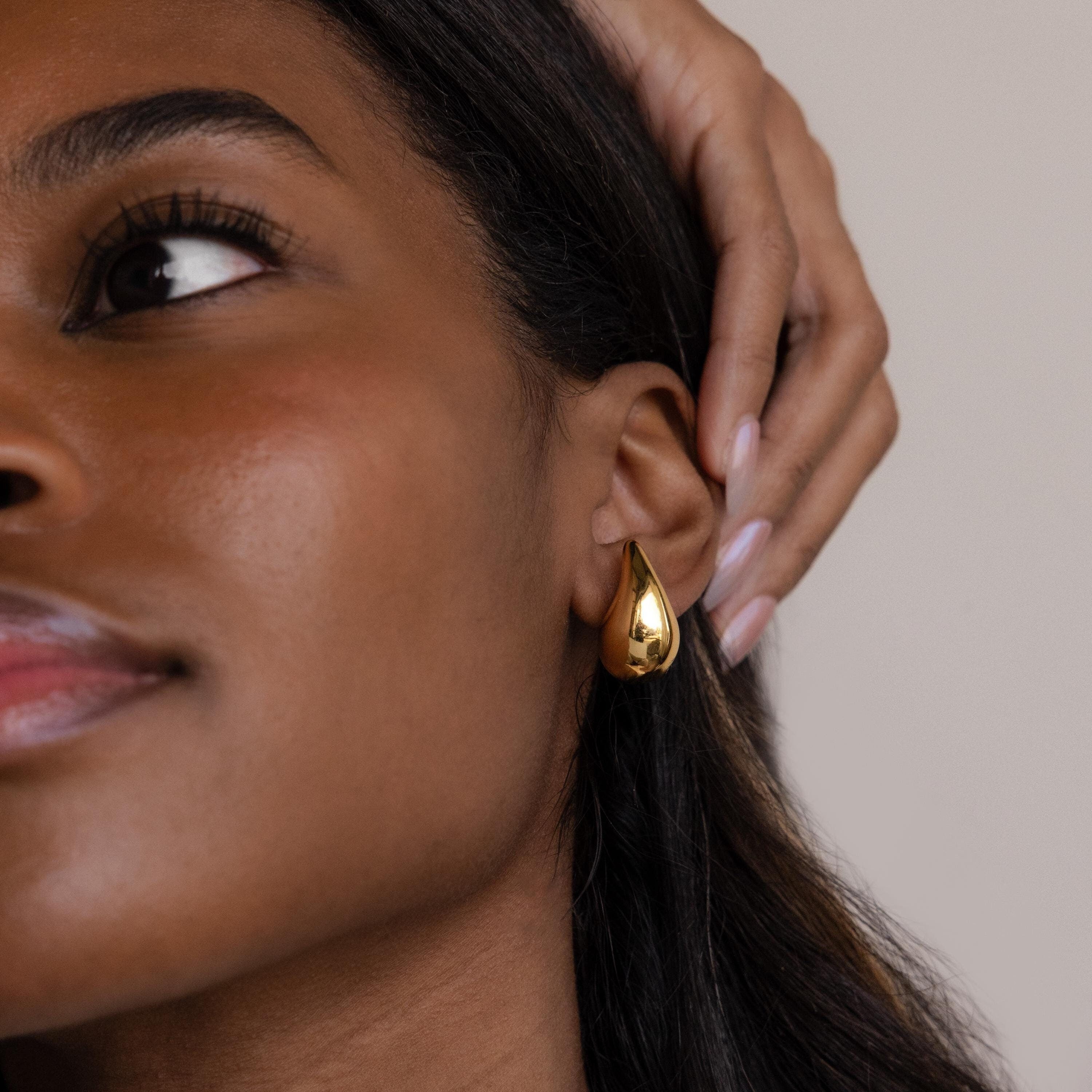 A woman touches her ear, showcasing the Chunky Tear Drop Studs—shiny, oversized stud earrings—highlighted in a close-up.
