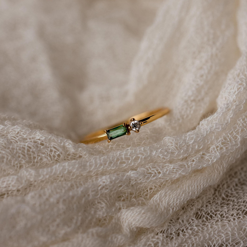 The Toi et Moi Baguette Birthstone Ring, with a green rectangular gem and a small clear stone, rests on cream-colored fabric.