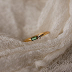 The Toi et Moi Baguette Birthstone Ring, with a green rectangular gem and a small clear stone, rests on cream-colored fabric.