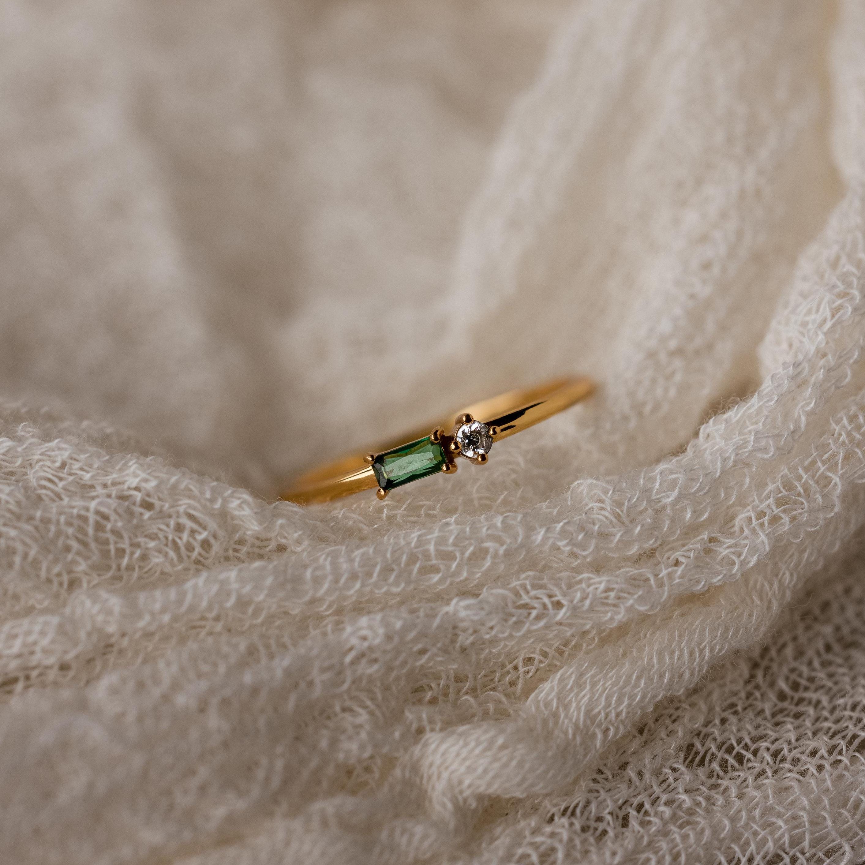 The Toi et Moi Baguette Birthstone Ring, with a green rectangular gem and a small clear stone, rests on cream-colored fabric.