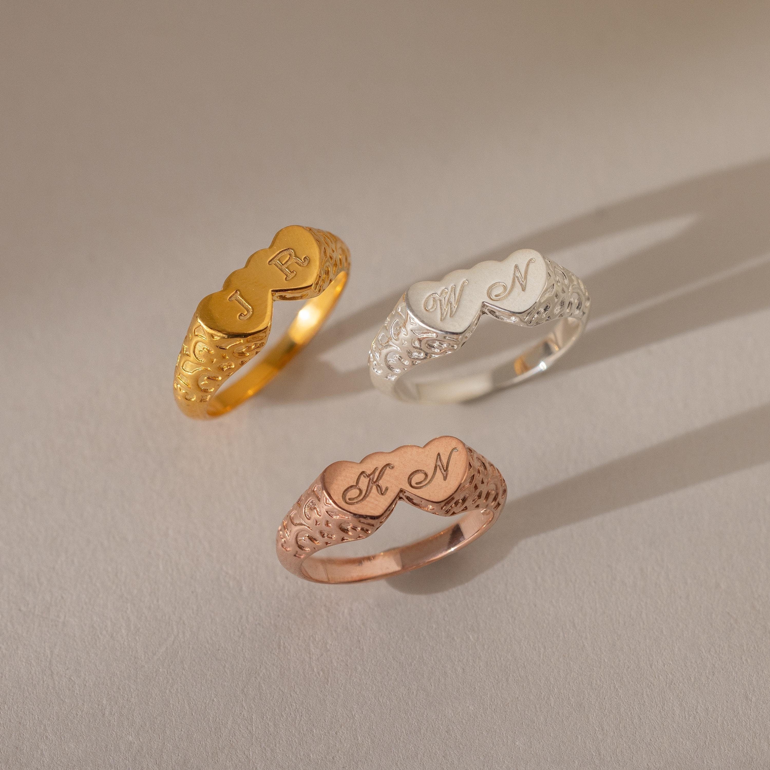 The Duo Initial Heart Ring features engraved decorative initials on a filigree band, available in gold, silver, and rose gold, displayed on a neutral background.