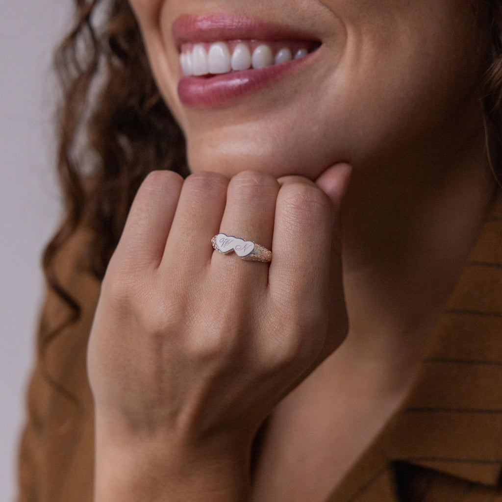 A woman smiles, displaying her Duo Initial Heart Ring with an engraved heart, stylishly paired with a brown pinstripe top.