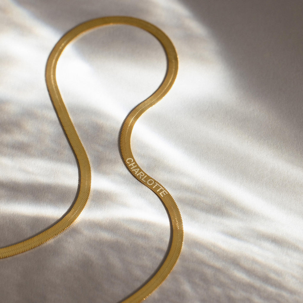 An Engraved Herringbone Name Necklace with CHARLOTTE rests on a softly lit, light-colored surface, radiating elegance and personal charm.