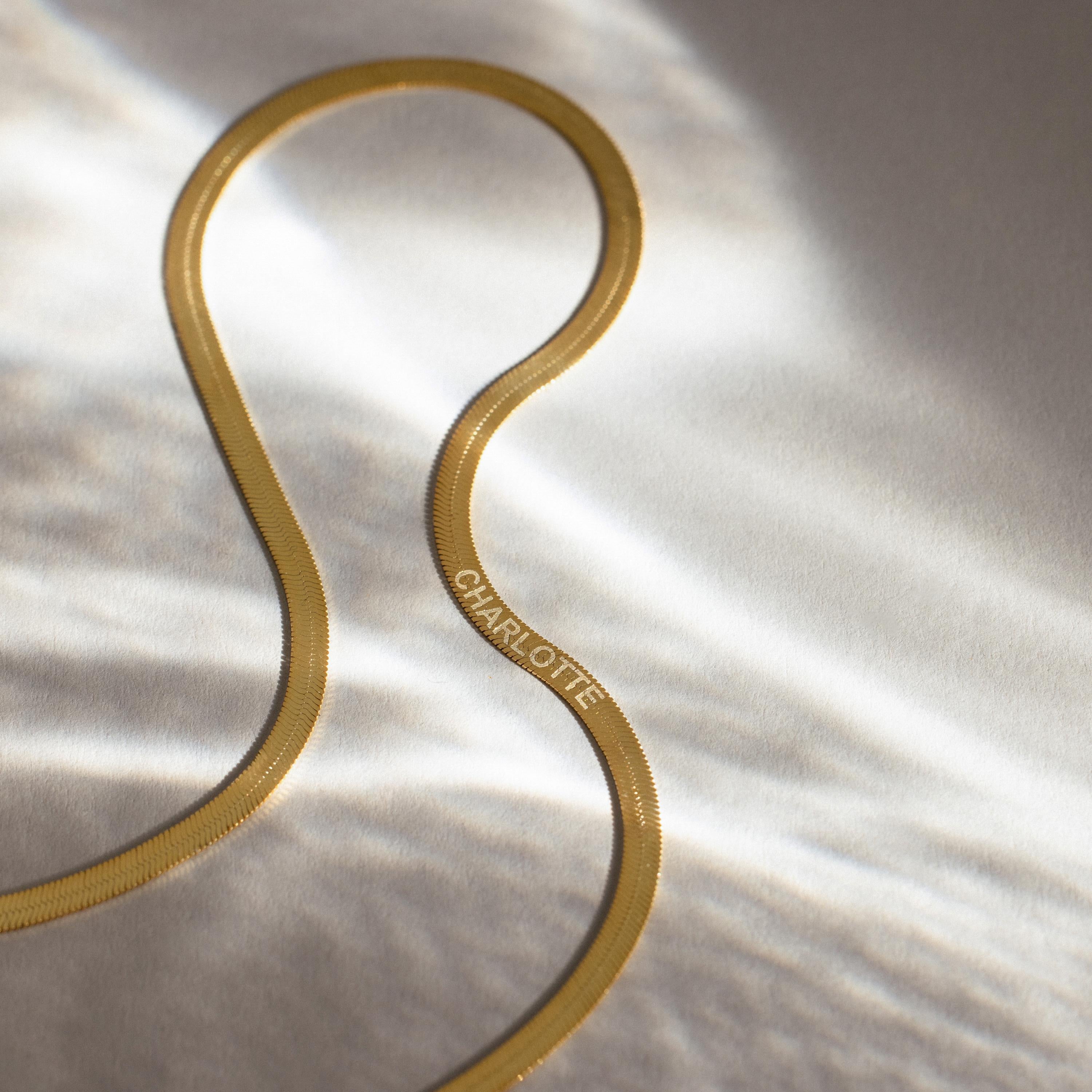 An Engraved Herringbone Name Necklace with CHARLOTTE rests on a softly lit, light-colored surface, radiating elegance and personal charm.