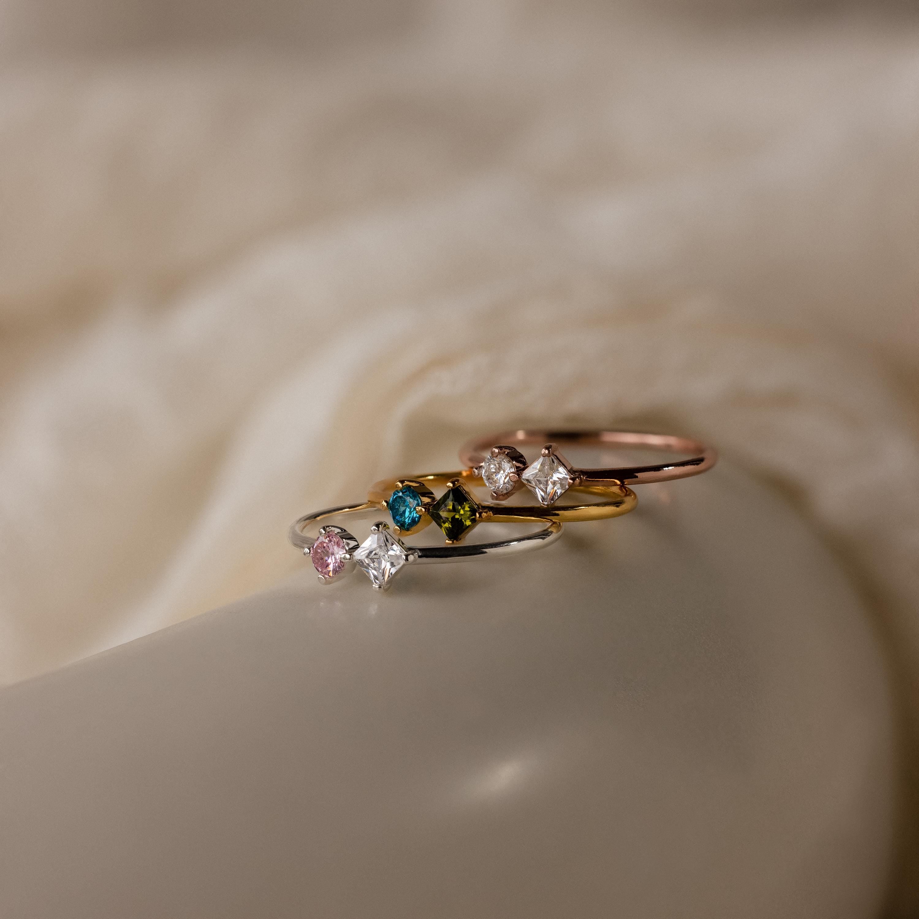 The Toi et Moi Princess Birthstone Ring features colorful gemstones set in gold, silver, and rose gold bands—ideal for creating a personalized or customized ring stack on soft, creamy fabric.
