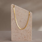 The Engraved Herringbone Name Necklace is displayed draped over a tall, angled beige stone block set against a neutral background.
