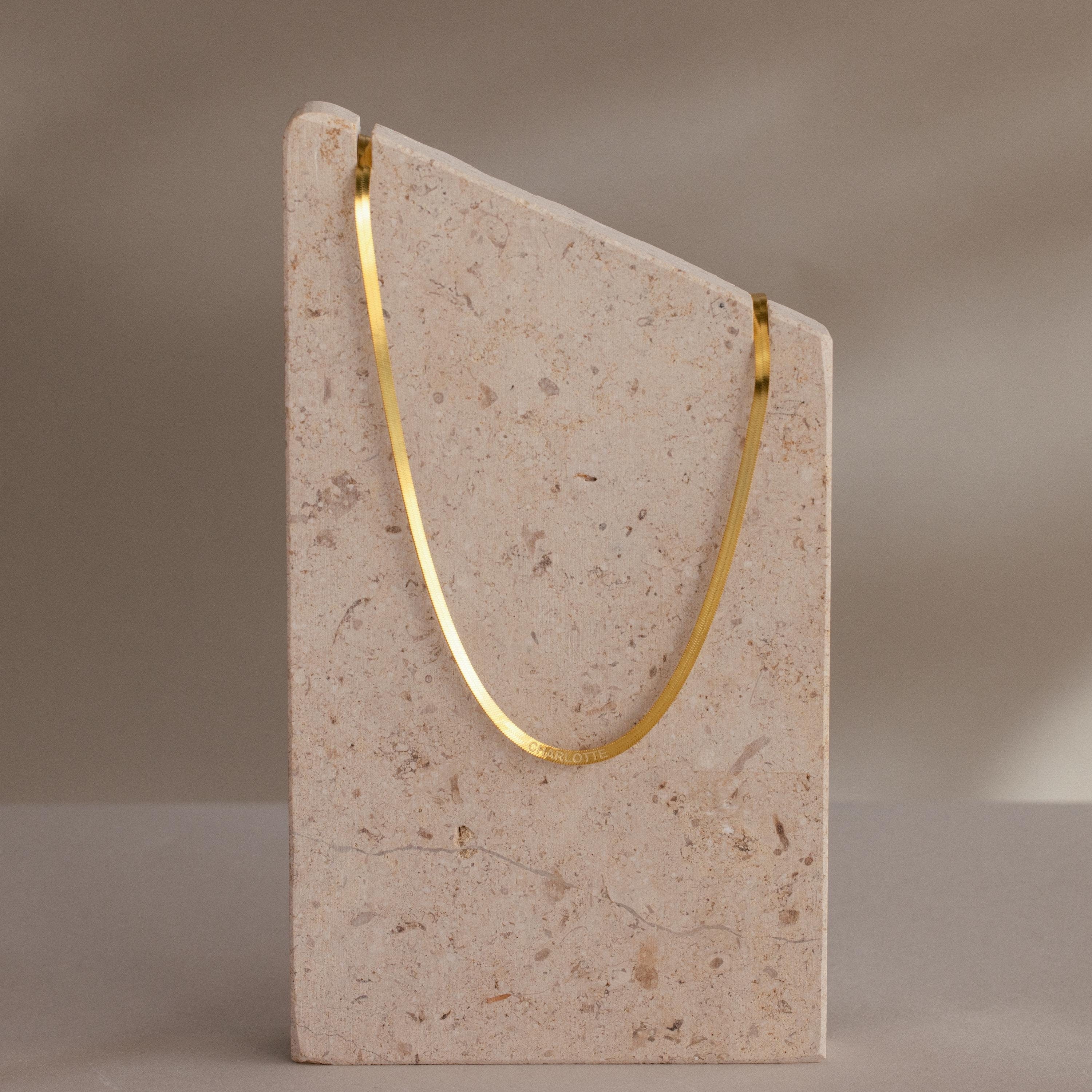 The Engraved Herringbone Name Necklace is displayed draped over a tall, angled beige stone block set against a neutral background.