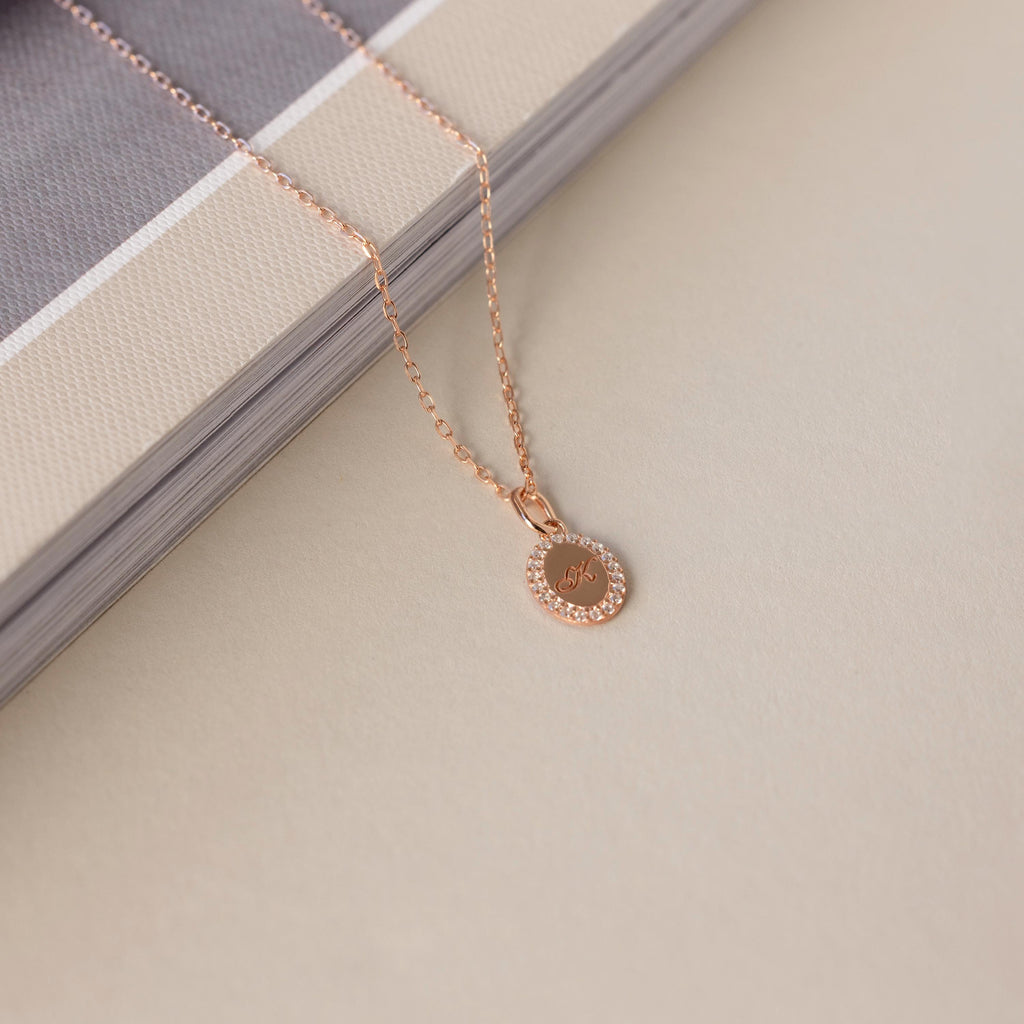 The Pave Initial Pendant Necklace in rose gold features a round pendant engraved with J, shown resting on a beige and gray book.