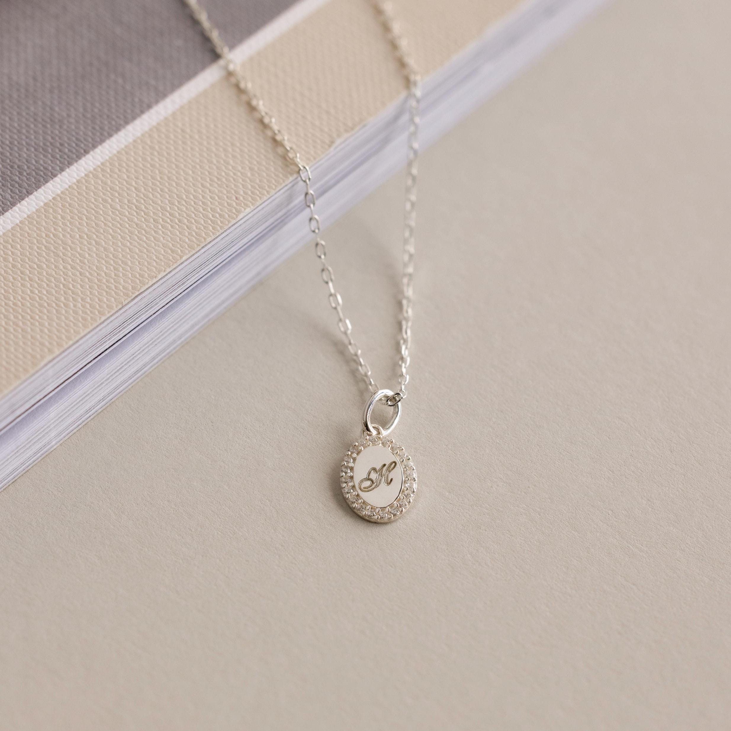 The Pave Initial Pendant Necklace with a round, diamond-accented K charm lies on a beige surface by a book, adding elegant style to this personalized jewelry display.
