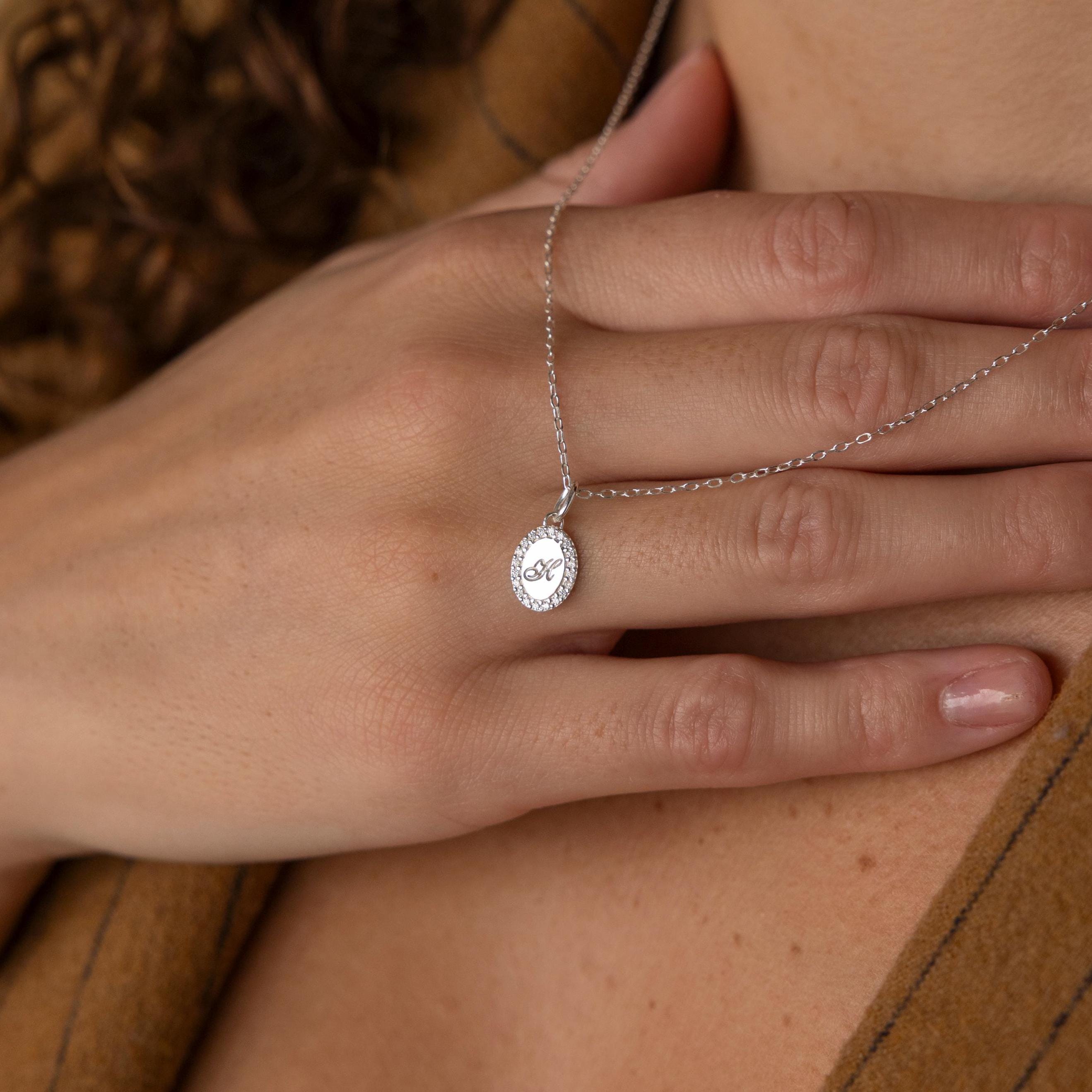 A hand touches the Pave Initial Pendant Necklace—a personalized silver oval pendant with an engraved initial—worn gracefully around the neck.