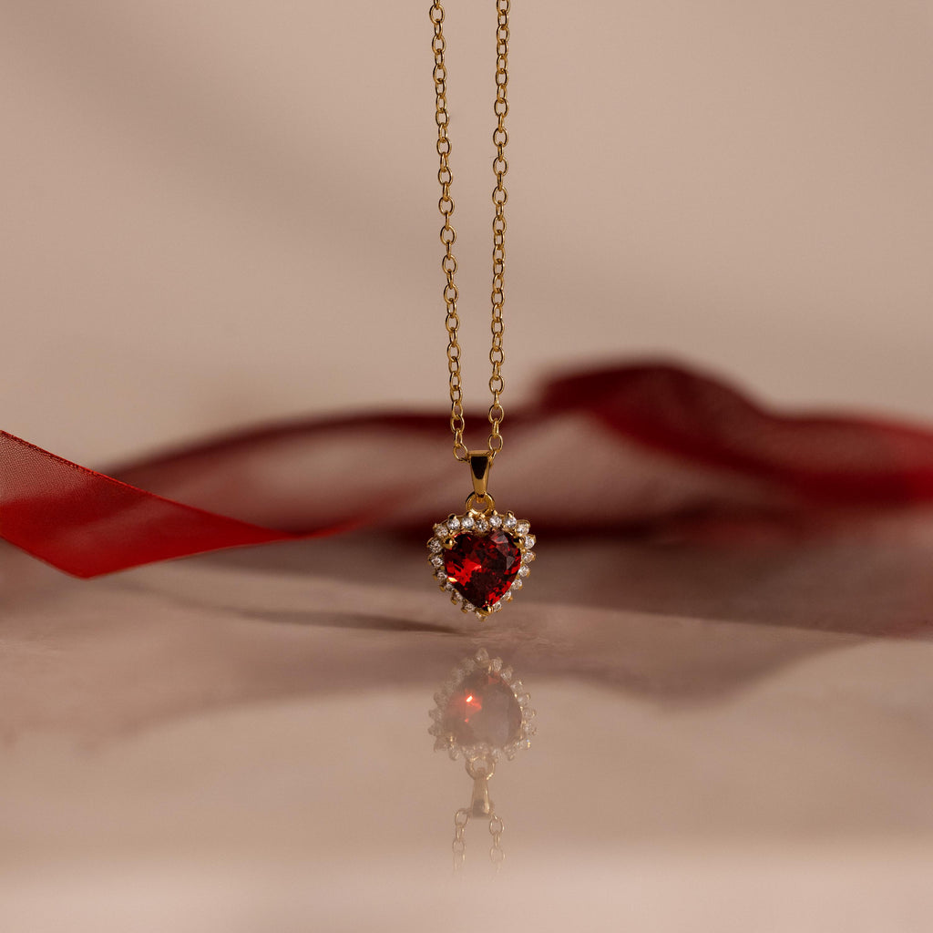 The Pave Garnet Heart Necklace features a heart-shaped red garnet pendant accented with sparkling gems, beautifully displayed on a reflective surface—a stunning birthstone necklace for any occasion.