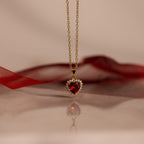 The Pave Garnet Heart Necklace features a heart-shaped red garnet pendant accented with sparkling gems, beautifully displayed on a reflective surface—a stunning birthstone necklace for any occasion.