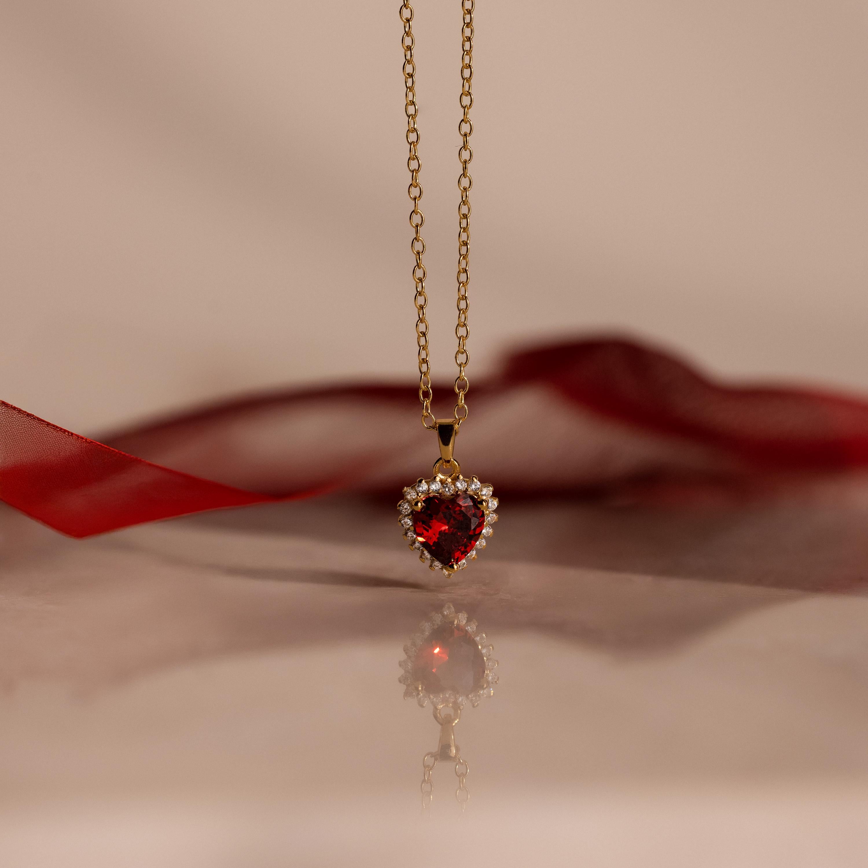 The Pave Garnet Heart Necklace features a heart-shaped red garnet pendant accented with sparkling gems, beautifully displayed on a reflective surface—a stunning birthstone necklace for any occasion.