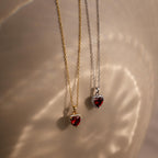 Two Pave Garnet Heart Necklaces with heart-shaped red gemstones, one on a gold chain and another on a silver chain, brilliantly illuminated by soft lighting.
