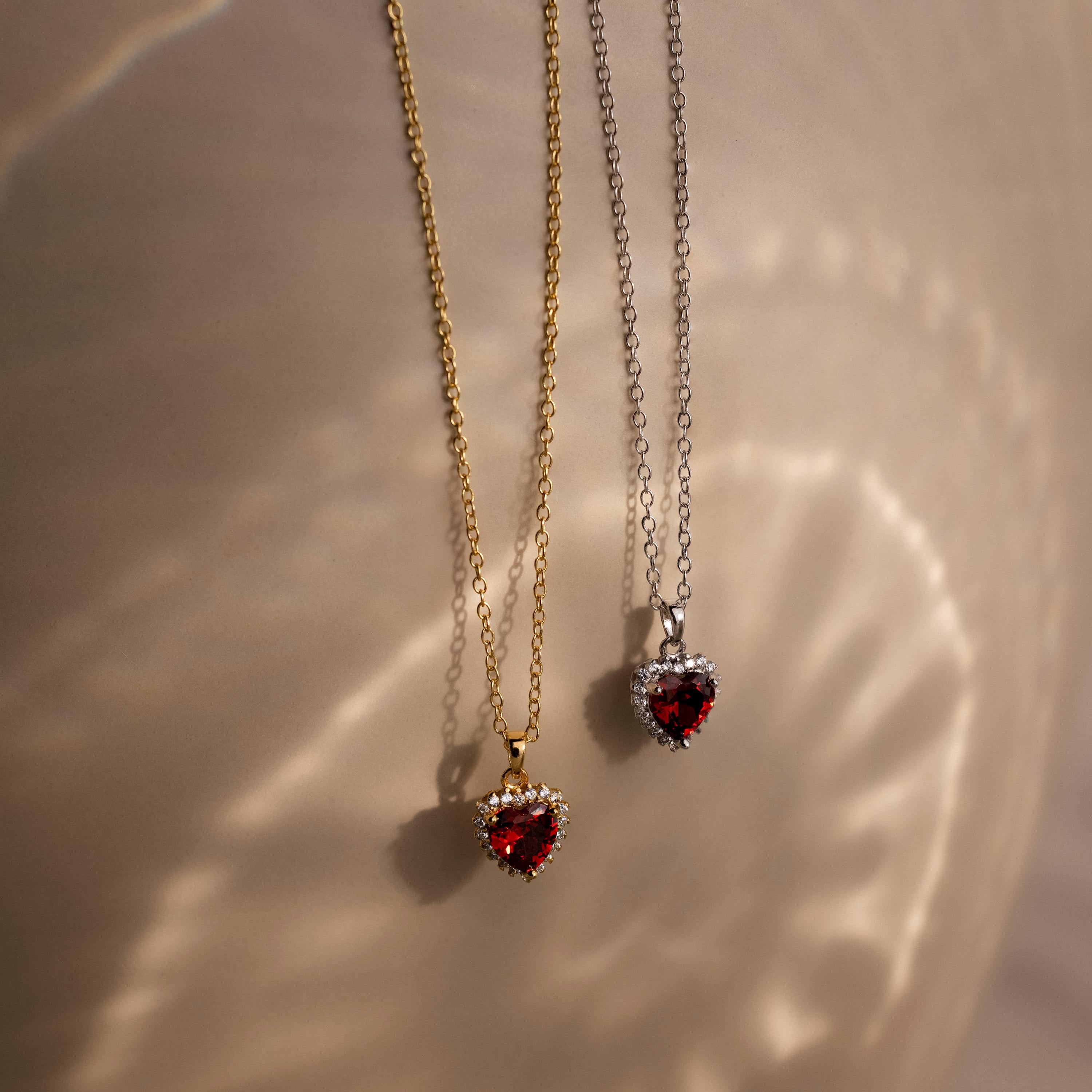 Two Pave Garnet Heart Necklaces with heart-shaped red gemstones, one on a gold chain and another on a silver chain, brilliantly illuminated by soft lighting.