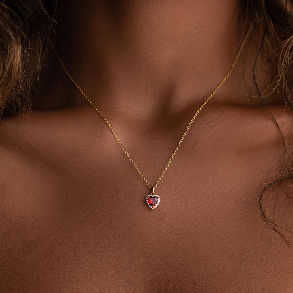 The Pave Garnet Heart Necklace, featuring a gold chain and a red heart-shaped garnet pendant surrounded by sparkling diamonds, adorns a woman's neck.