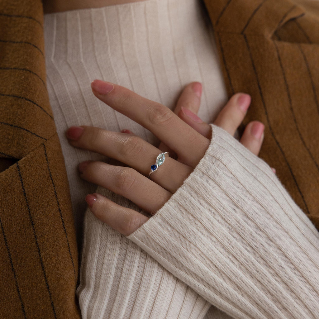 A hand adorned with the Toi et Moi Infinity Birthstone Ring rests on a chest, complemented by a cream ribbed sweater and brown pinstripe blazer—a subtle yet striking symbol of love.