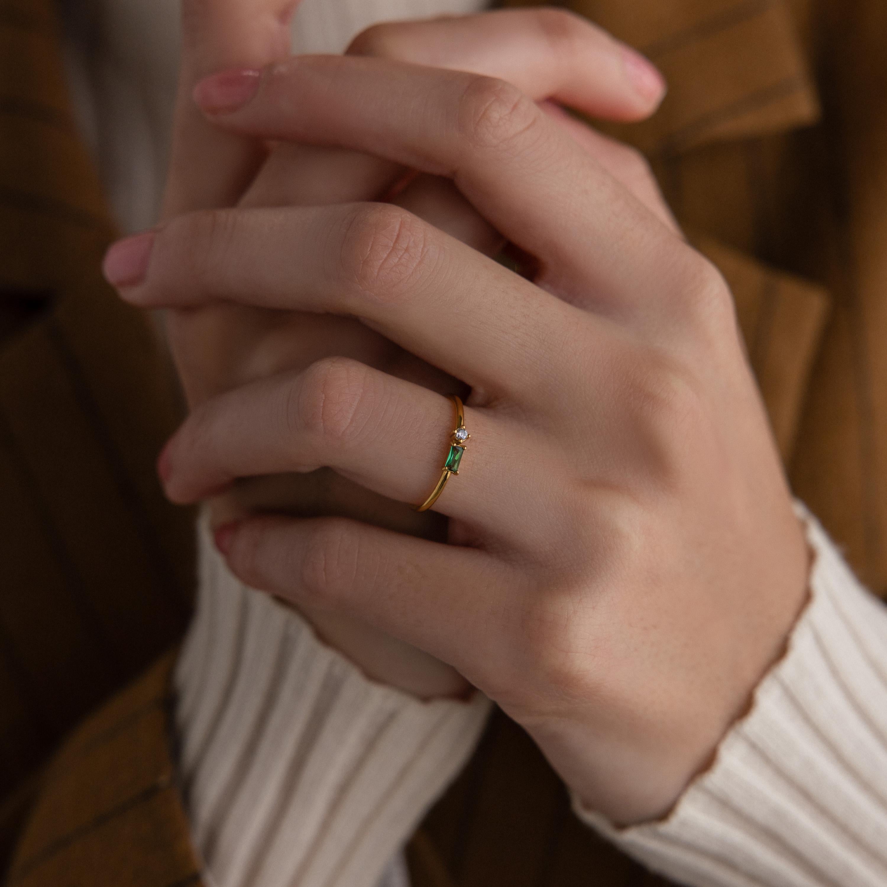 A hand wearing the Toi et Moi Baguette Birthstone Ring with a green stone, fingers gently clasped, in a striped brown jacket and cream sweater.