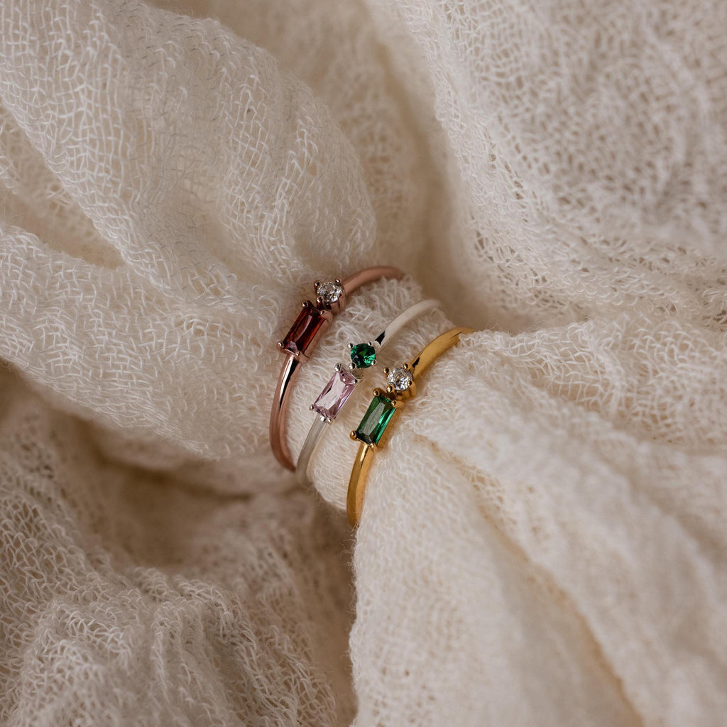 The Toi et Moi Baguette Birthstone Ring features sparkling colored gemstones and diamonds, elegantly displayed on textured cream fabric.