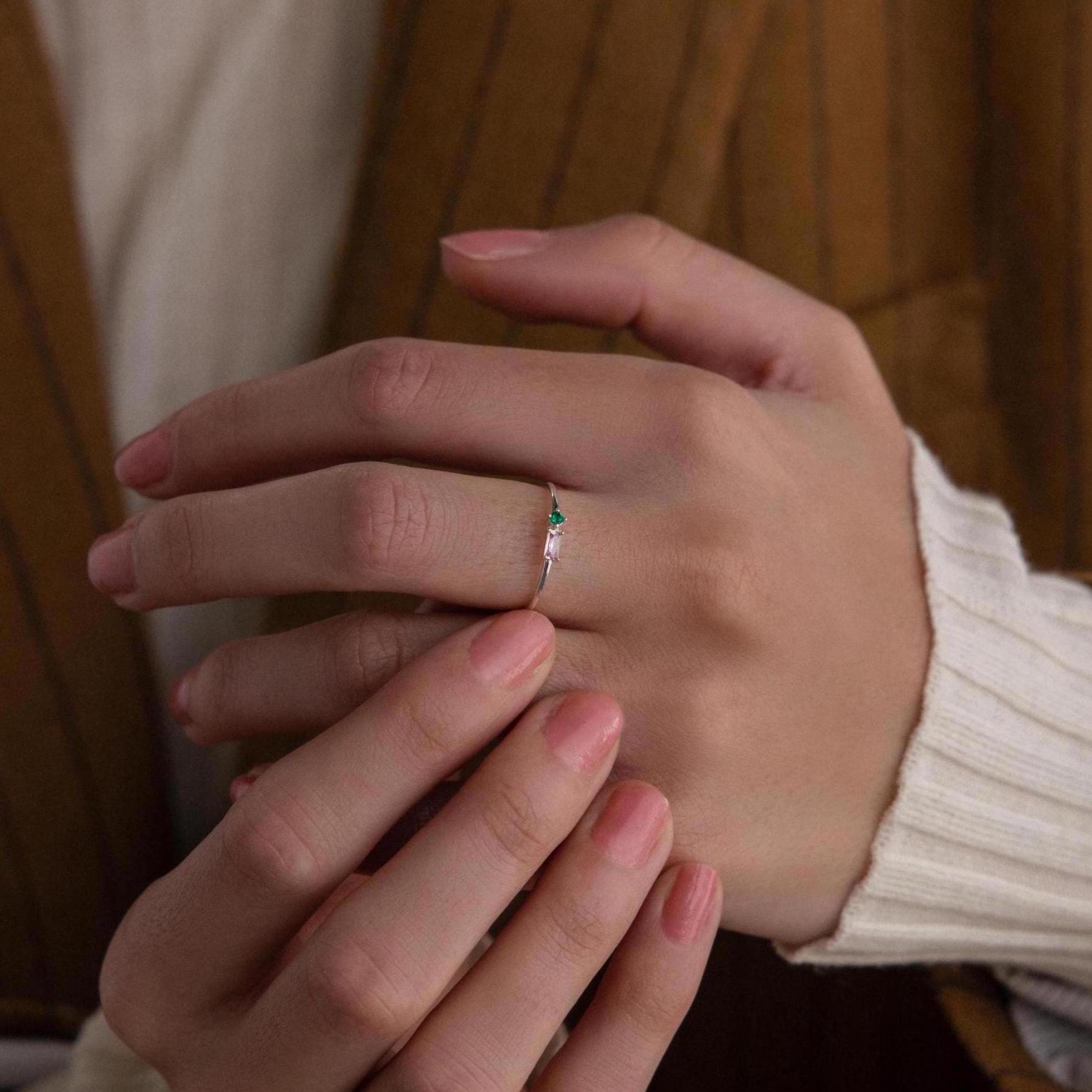A hand adorned with the Toi et Moi Baguette Birthstone Ring, featuring a delicate, personalized design and a small green gemstone, with fingers gently touching the ring.