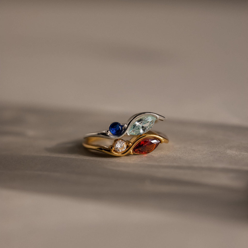 The Toi et Moi Infinity Birthstone Ring, featuring blue, green, red, and clear personalized gemstones, glimmers on a neutral background—a distinctive symbol of love.