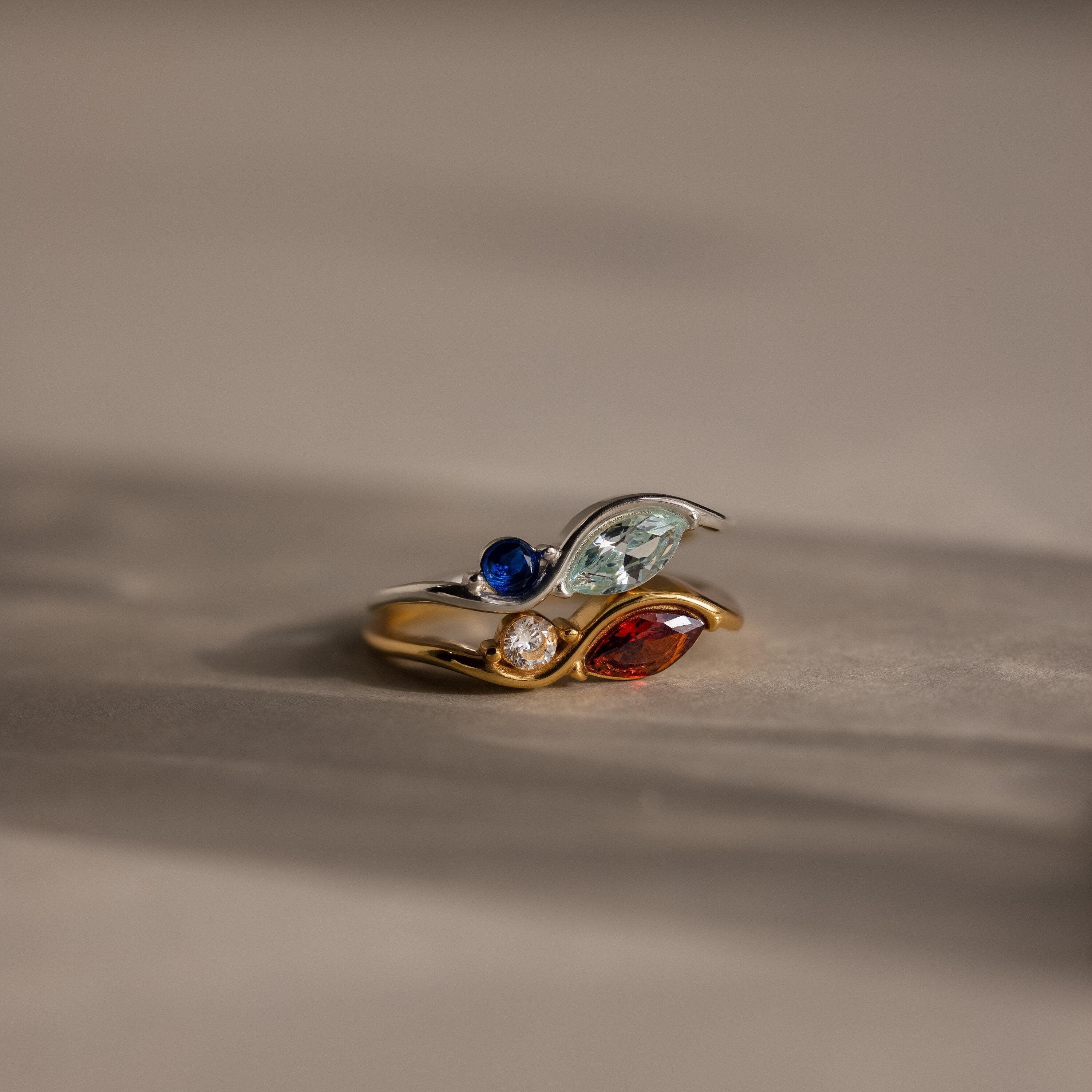 The Toi et Moi Infinity Birthstone Ring, featuring blue, green, red, and clear personalized gemstones, glimmers on a neutral background—a distinctive symbol of love.