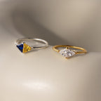 A Toi et Moi Triangle Birthstone Ring in silver with blue and yellow stones, paired with a gold band featuring a clear white gem, are displayed on a neutral surface.