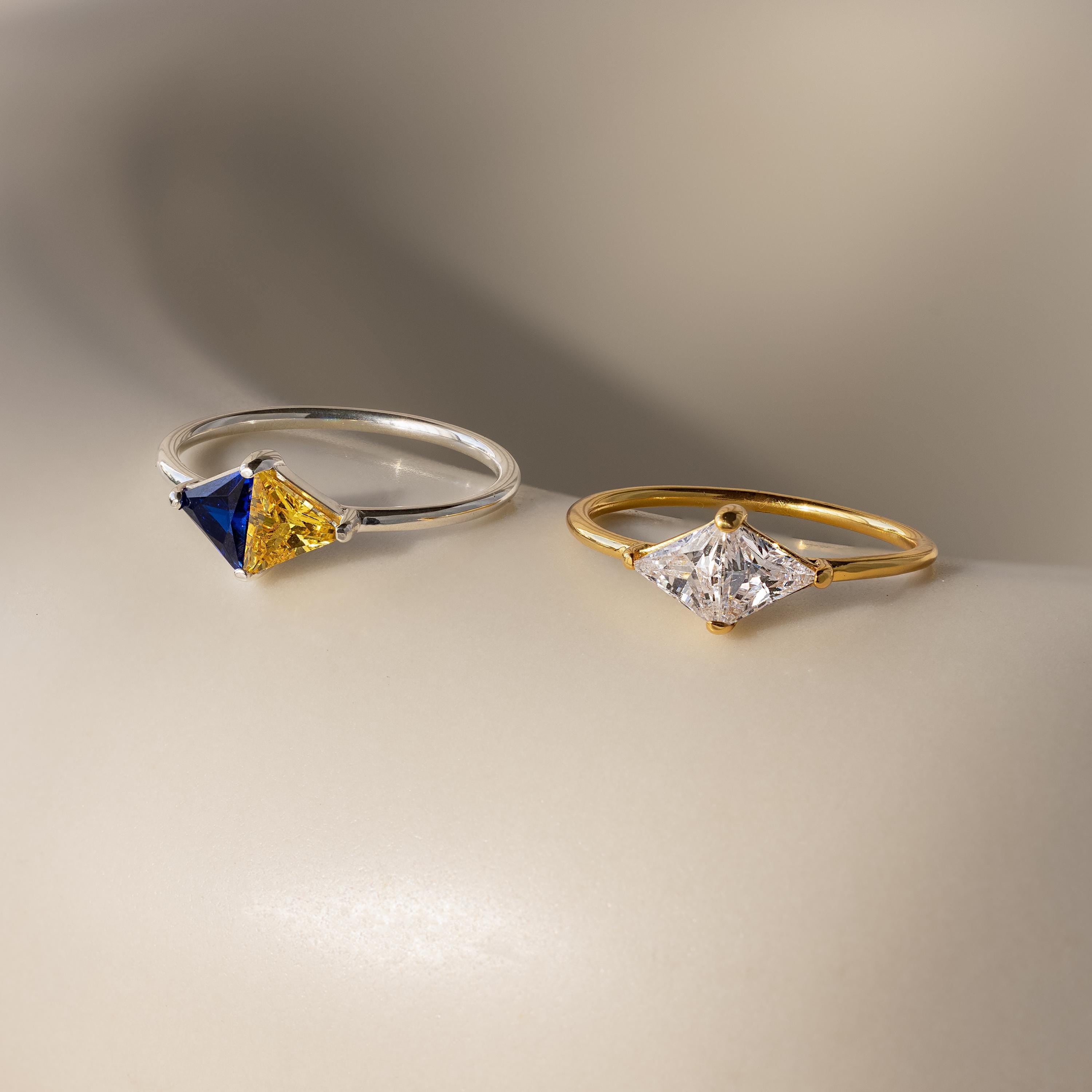 A Toi et Moi Triangle Birthstone Ring in silver with blue and yellow stones, paired with a gold band featuring a clear white gem, are displayed on a neutral surface.