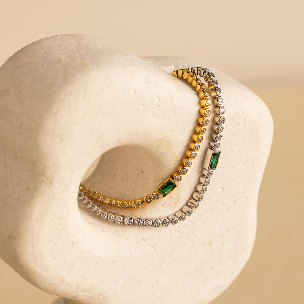 An elegant Emerald Baguette Tennis Bracelet with green gemstones drapes over a smooth, sculptural stone—perfect for lovers of statement jewelry.