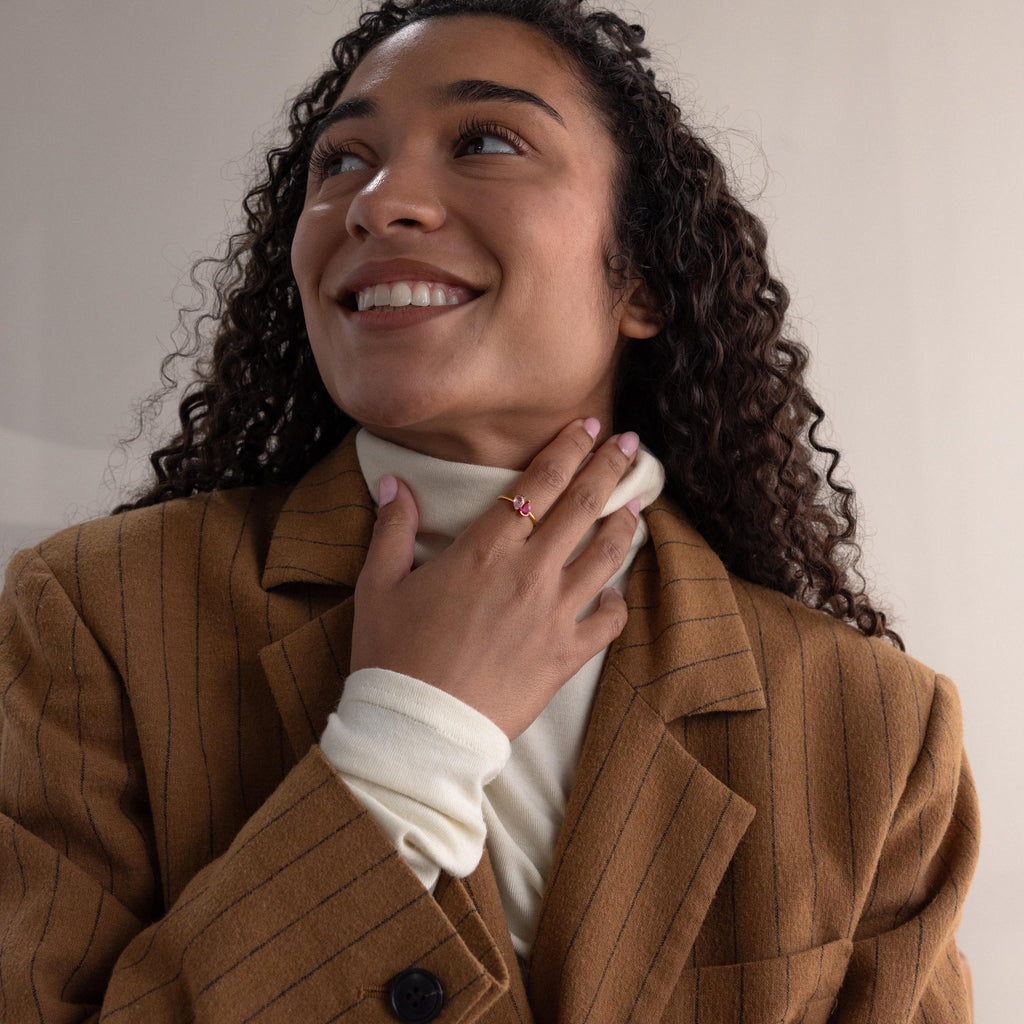 Smiling woman in a brown pinstripe blazer touches her neck while wearing the Toi et Moi Pear Birthstone Ring—a perfect romantic gift.