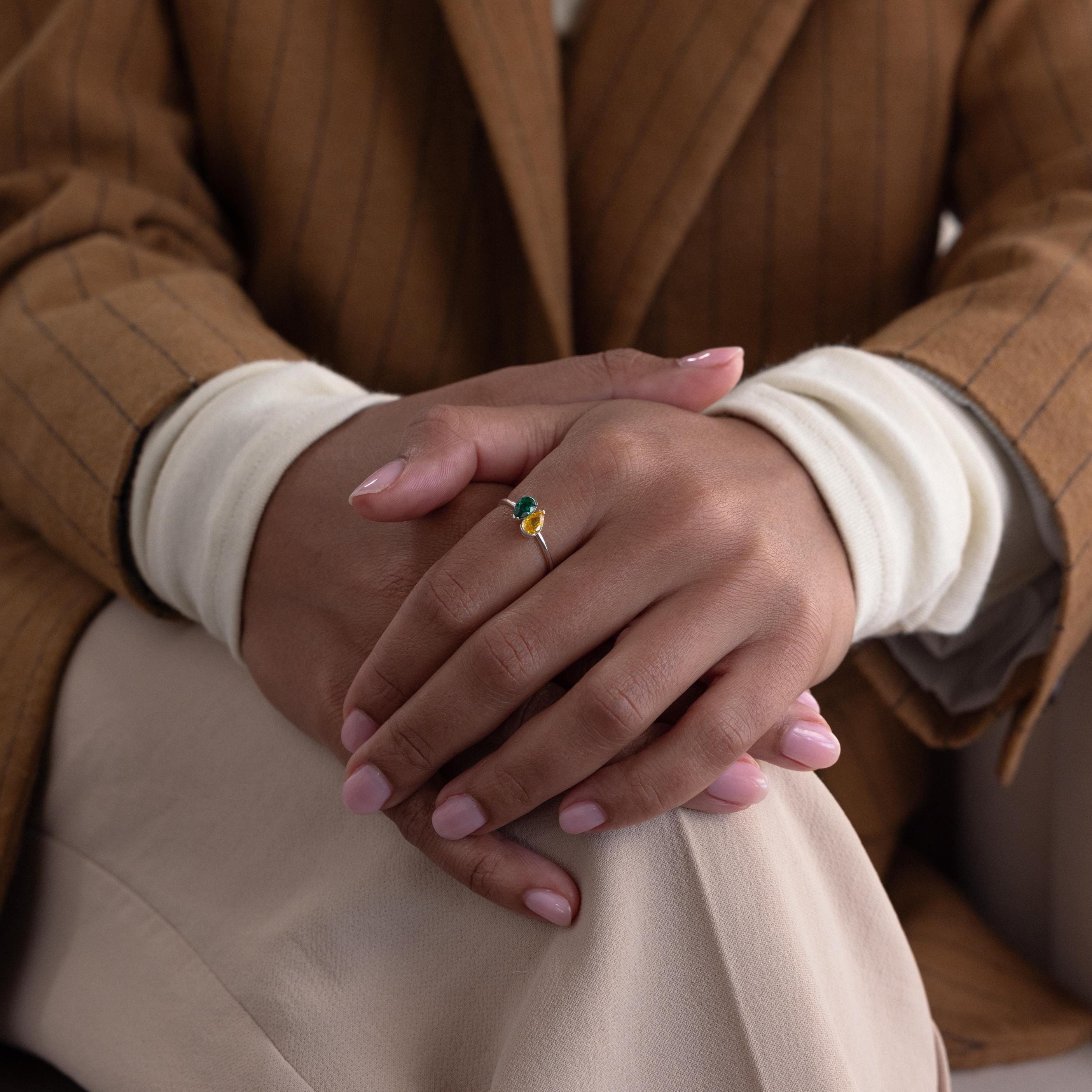 A person in a brown pinstripe suit jacket with hands folded showcases the Toi et Moi Pear Birthstone Ring with a green stone—an elegant and unique romantic gift.