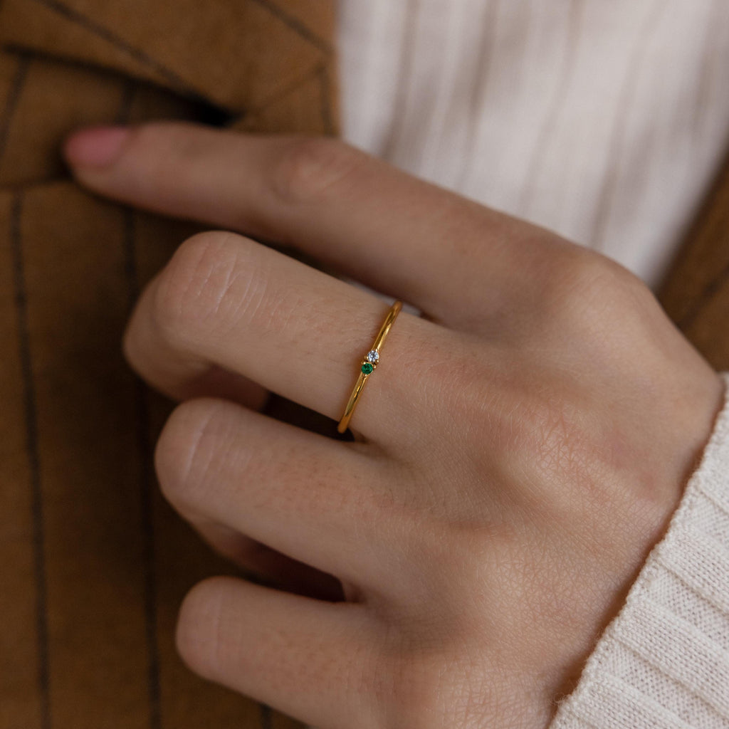 A hand wearing the Mini Toi et Moi Birthstone Ring, a delicate gold band with a small green gemstone, shown against a brown jacket and cream sweater.