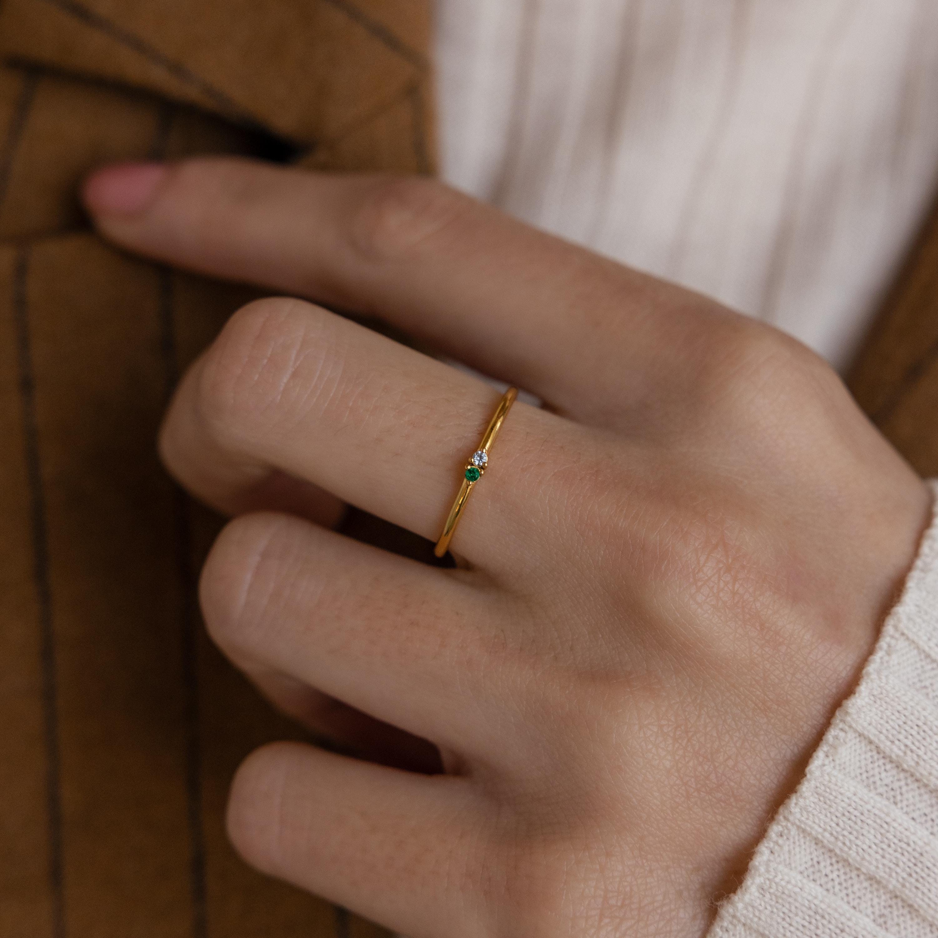 A hand wearing the Mini Toi et Moi Birthstone Ring, a delicate gold band with a small green gemstone, shown against a brown jacket and cream sweater.