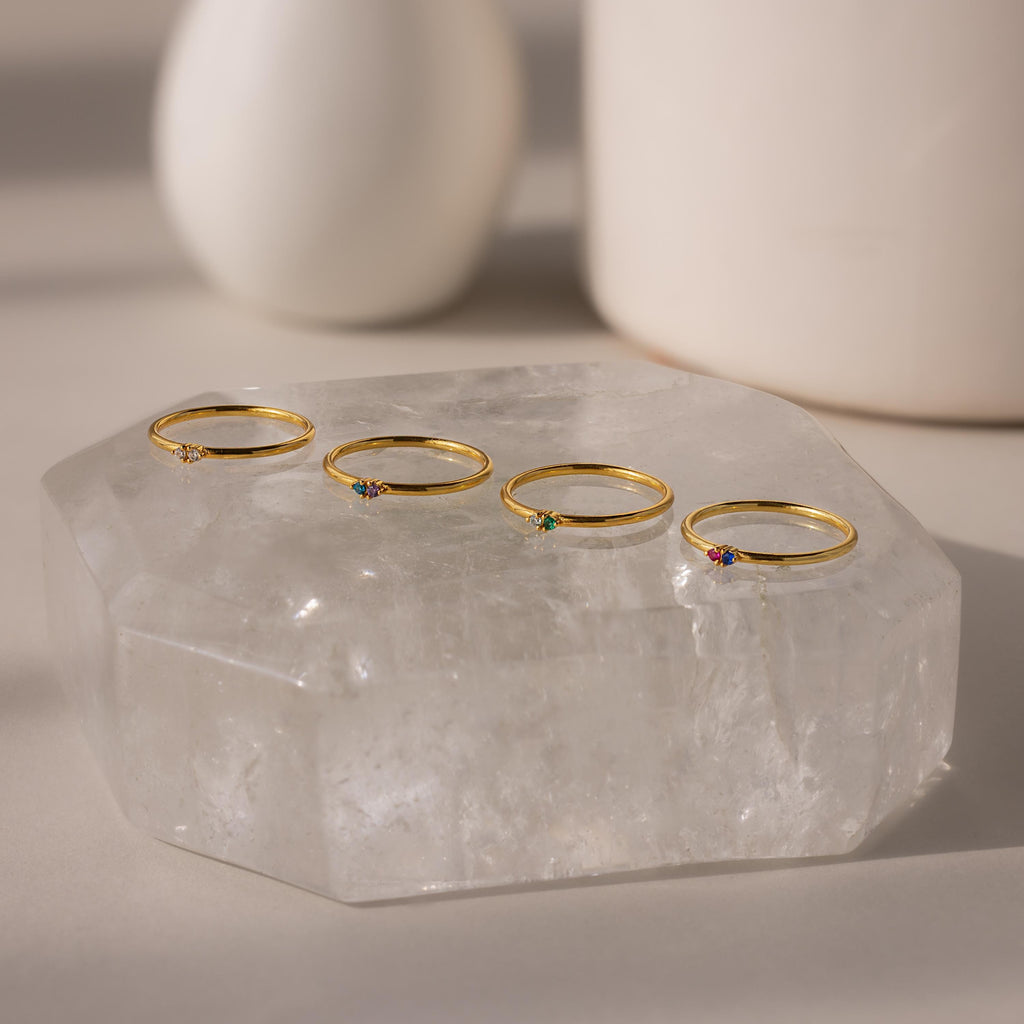 Four delicate gold rings, featuring the Mini Toi et Moi Birthstone Ring, display colorful gemstones and sparkle on a large clear crystal slab.