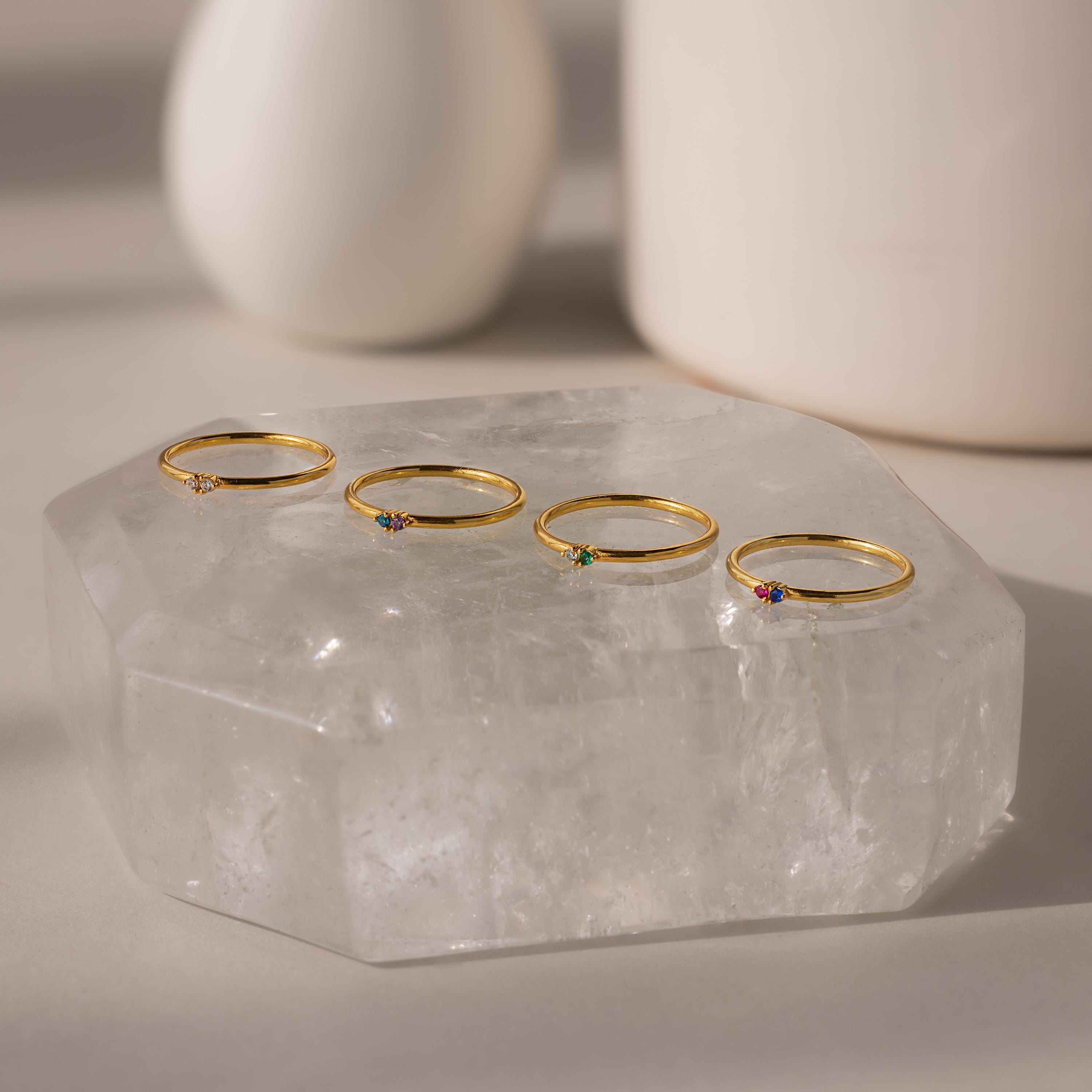 Four delicate gold rings, featuring the Mini Toi et Moi Birthstone Ring, display colorful gemstones and sparkle on a large clear crystal slab.