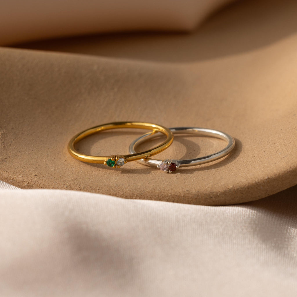 Two delicate rings—a gold Mini Toi et Moi Birthstone Ring with a green birthstone and a silver ring with a red stone—rest gracefully on a soft beige surface.